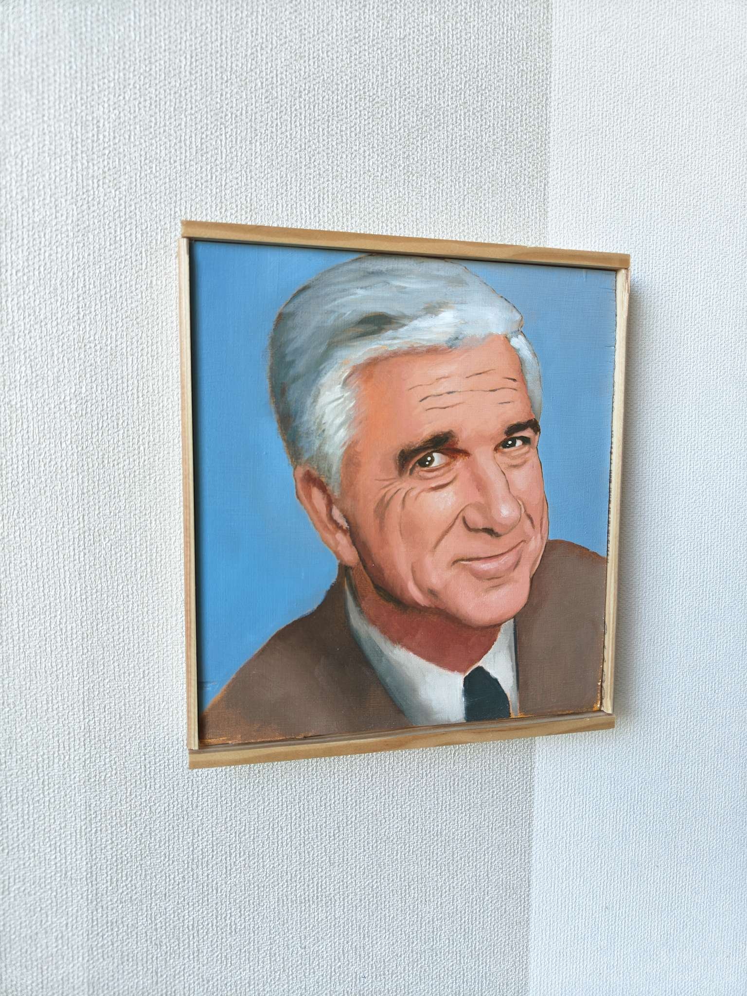 Vasemmalta viistosta kuvattu Oskari Myllymäen alkuperäinen öljyvärimaalaus, joka esittää näyttelijä Leslie Nielseniä Frank Drebinin roolissa. Teos on maalattu puupaneelille ja siinä on vaalea puukehys.