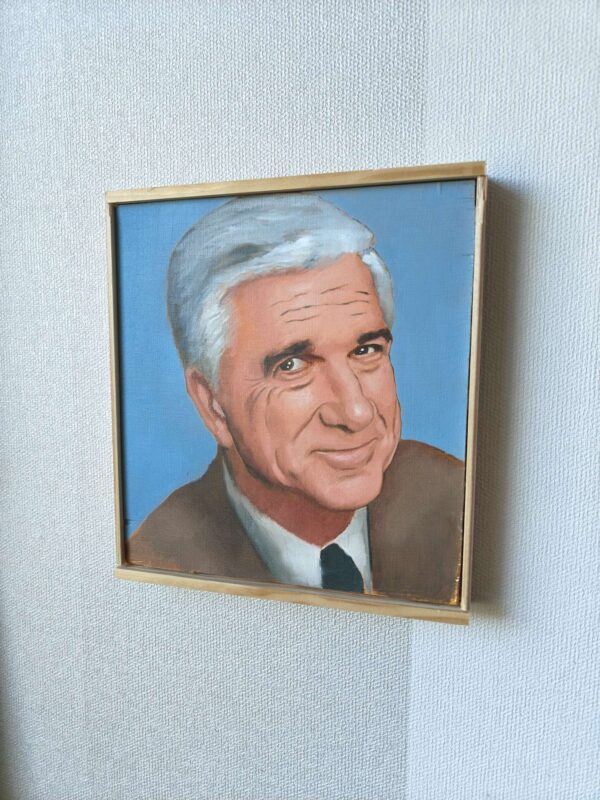 Lähikuva Oskari Myllymäen maalaamasta Frank Drebin (Leslie Nielsen) -öljyvärimaalauksesta, tuoden esiin kasvojen tarkat yksityiskohdat ja elokuvallisen realismin.